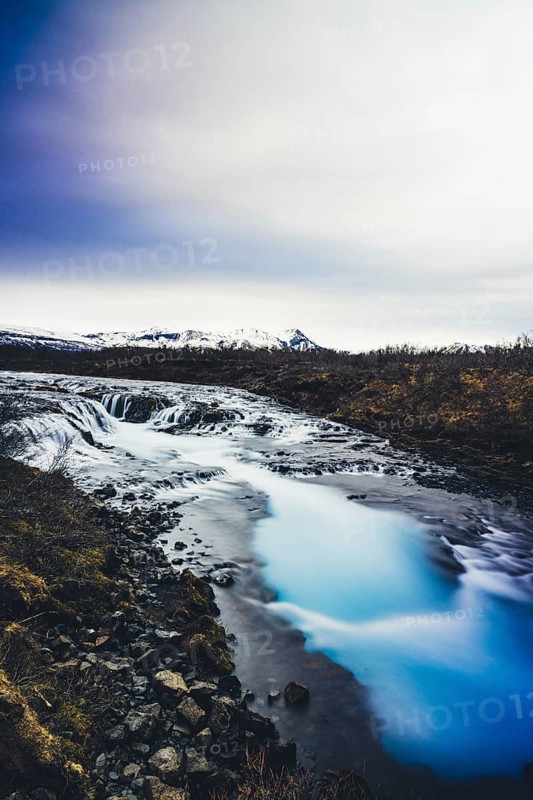 The stunning Bruarfoss waterfall, known for its crystal-clear turquoise waters, flowing over dark volcanic rocks surrounded by Iceland's rugged landscape and overcast skies