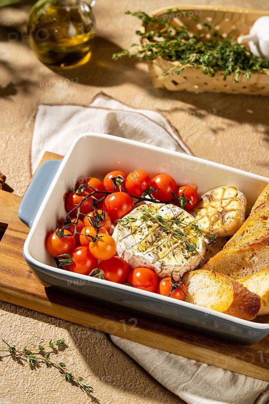 A dish featuring baked Camembert cheese, surrounded by cherry tomatoes and garlic, served with crusty bread. Garnished with herbs for a flavorful presentation