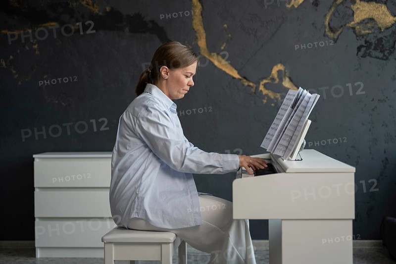 Woman dressed in white plays the piano in a stylish room with dark, textured walls and gold accents. Sheet music rests on the instrument as she focuses on her practice
