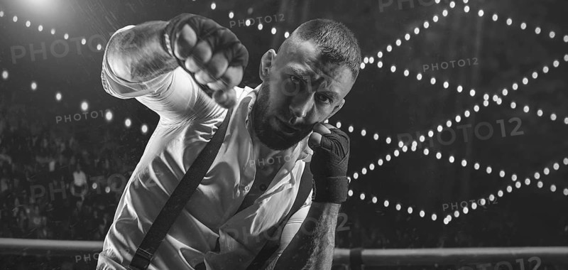 Black and white image of a bearded man in a white shirt and suspenders in the ring. The concept of fist underground fights.