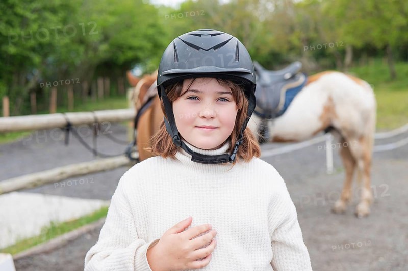 A young girl wearing a helmet enjoys a sunny day on the farm, standing beside her horse. The lush green surroundings add to the idyllic outdoor experience