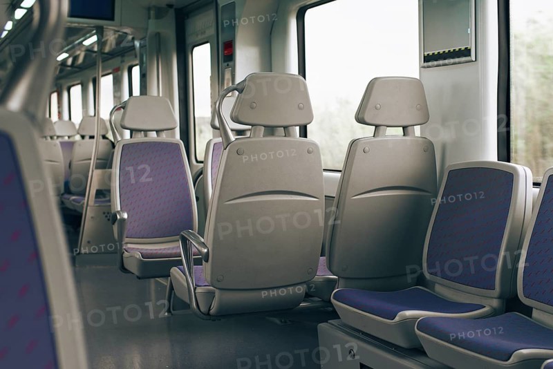 The photo depicts an empty train interior featuring modern gray seats with purple fabric accents. Sunlight filters through large windows, creating a calm and serene atmosphere