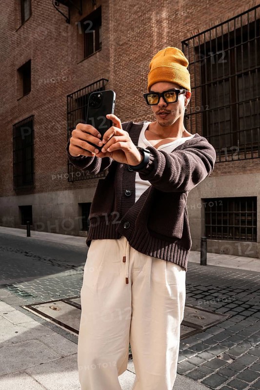 A fashionable latin man in a yellow beanie, sunglasses, and casual attire takes a selfie with a smartphone in an urban setting, showcasing a modern and trendy style