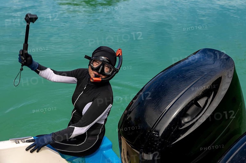 A snorkeler in a wetsuit and diving gear sits on a boat, holding an action camera, prepared for an underwater exploration in clear blue waters of Indonesia