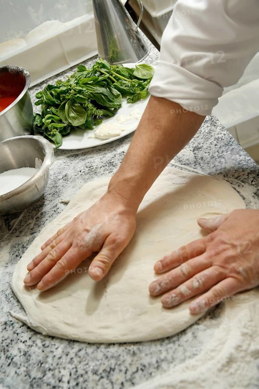 Cropped unrecognizable chef skillfully prepares pizza dough on a floured surface in a kitchen, surrounded by fresh ingredients like basil and mozzarella