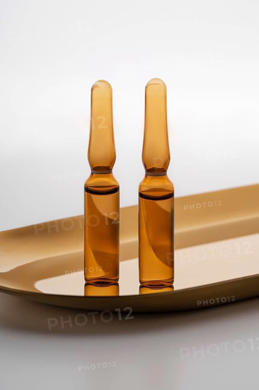 Close-up view of two amber-colored cosmetic ampoules containing anti-aging serum, displayed elegantly on a golden tray against a soft white background. Ideal for facial skincare routines