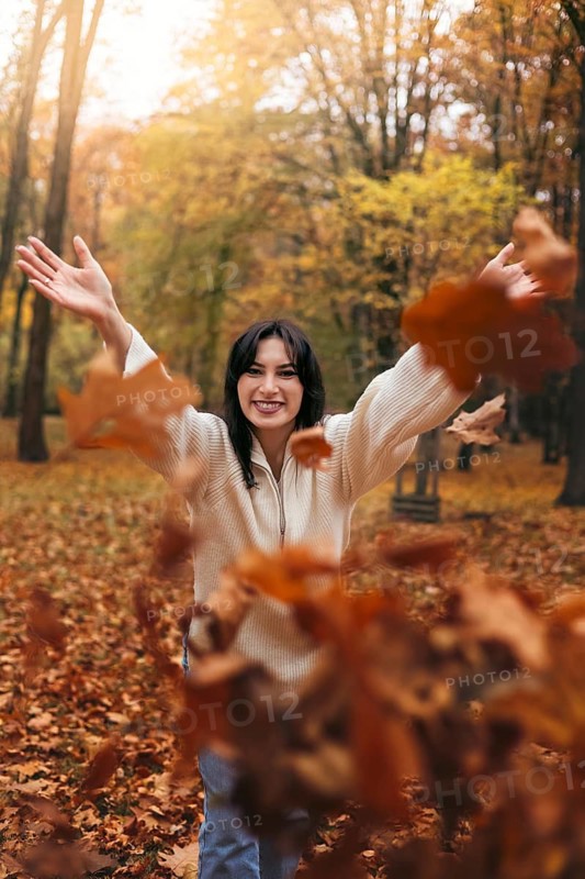 A joyful woman in a cozy sweater, standing amidst falling autumn leaves in a forest. The warm, golden tones of the leaves create a peaceful and cheerful atmosphere