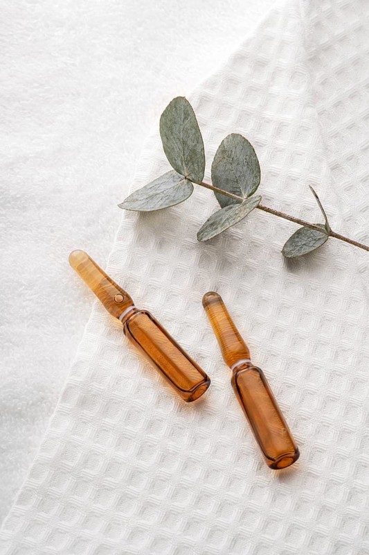 Elegant glass ampoules filled with anti-aging serum for face care, displayed on a textured white towel alongside a eucalyptus branch
