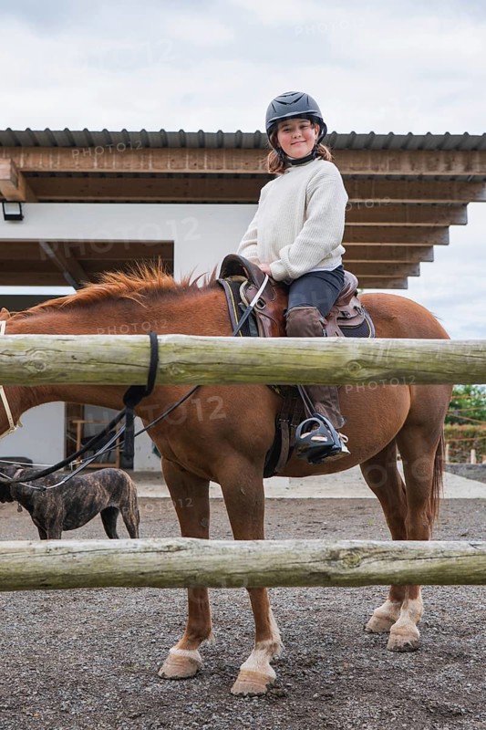 A young girl wearing a helmet enjoys a horseback ride at a farm. The scenery includes a stable, showcasing a delightful and adventurous day in nature