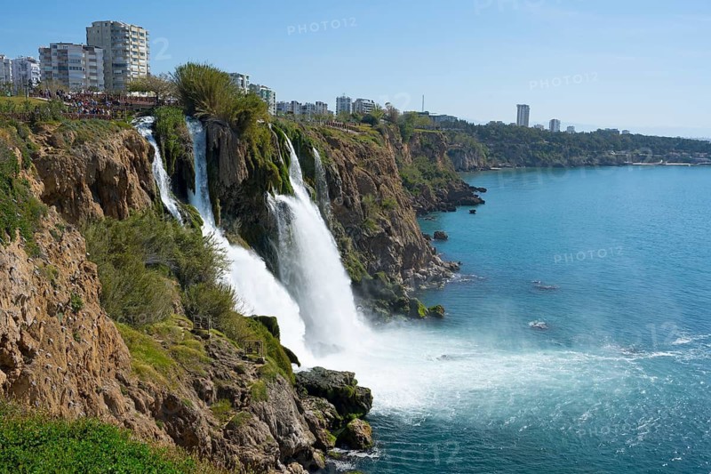 A waterfall falls from cliffs into the sea surrounded by buildings in a coastal town, Lower Düden Waterfall, Düden River, Antalya, Turkey