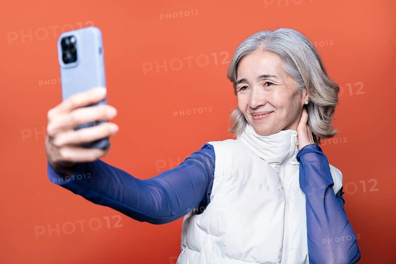 Asian senior woman with short gray hair takes a selfie with her smartphone against an orange background. She wears a white jacket and blue sleeves, smiling warmly