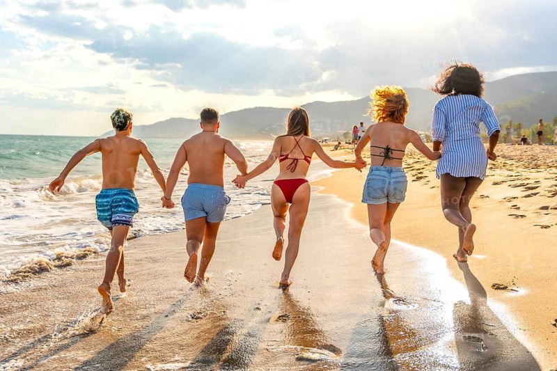 Five multi ethnic friends running on the beach at sunset holding hands, enjoying summer vacation together