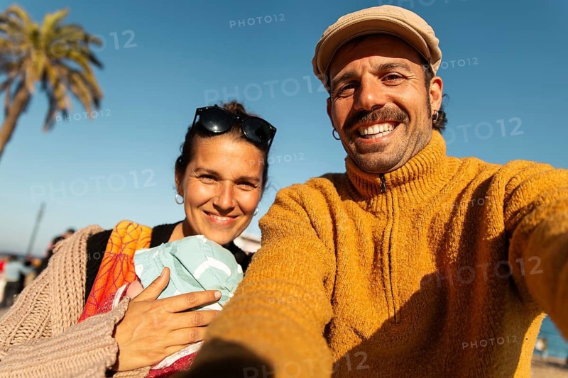 A cheerful couple takes a selfie with their baby on a sunny day in Barcelona. The man wears a mustard sweater and flat cap, while the woman cradles the baby against a vibrant backdrop