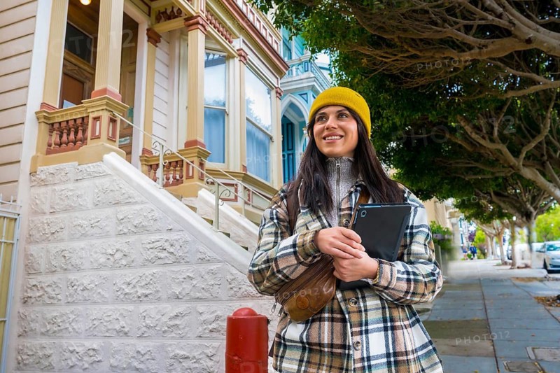 Young woman smiling, holding a tablet while walking on a sidewalk, enjoying urban tourism among historic victorian architecture and tree lined streets in san francisco