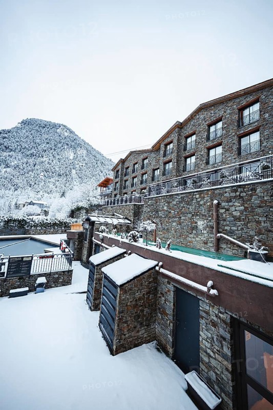 A serene winter scene showing a stone building nestled against a snow capped mountain in Andorra. Fresh snow blankets the structure and landscape, creating a tranquil, picturesque view