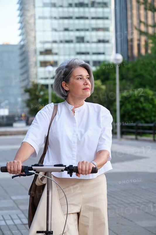 Mature woman with gray hair riding a scooter through a modern cityscape. She looks content and stylish, wearing a white blouse and beige pants. Background includes glass buildings and greenery