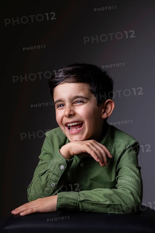 A portrait of a young boy wearing a green shirt, captured in a moment of joyful laughter against a neutral background. The image exudes happiness and lightheartedness
