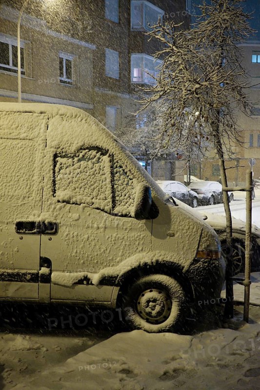 A van becomes blanketed in fresh snow at night, highlighting winter's impact on urban transport in the city