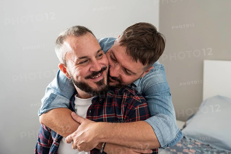 A joyful gay couple embraces lovingly in a cozy bedroom, exuding warmth and affection. One partner gently hugs the other from behind, both smiling brightly