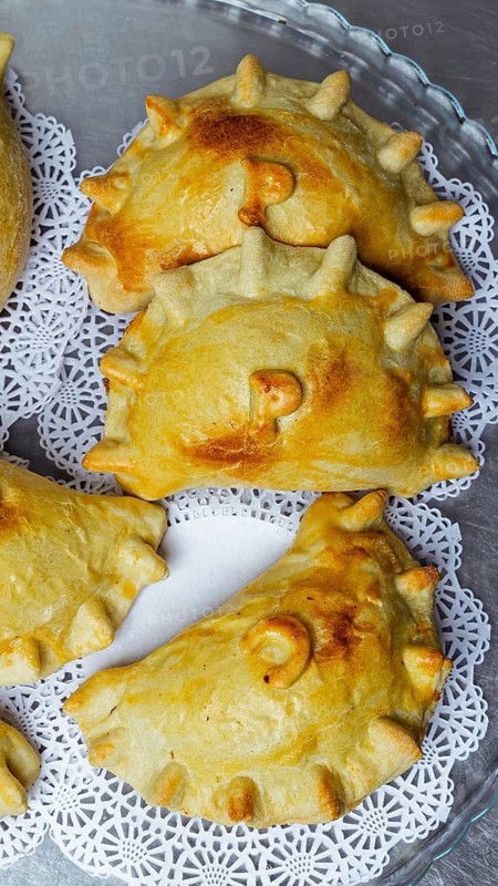 Close-up of golden brown, homemade baked empanadas with decorative edges and stamped tops, arranged on a tray lined with lace-patterned paper, showcasing traditional pastry detail