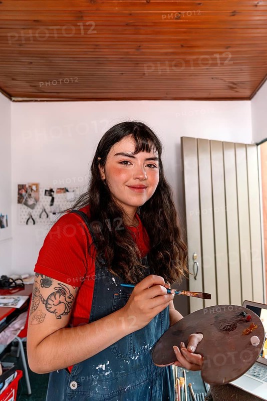 A young woman artist is painting a canvas in her home studio. She holds a palette and brush, surrounded by art supplies, radiating creativity and passion in an inspiring space