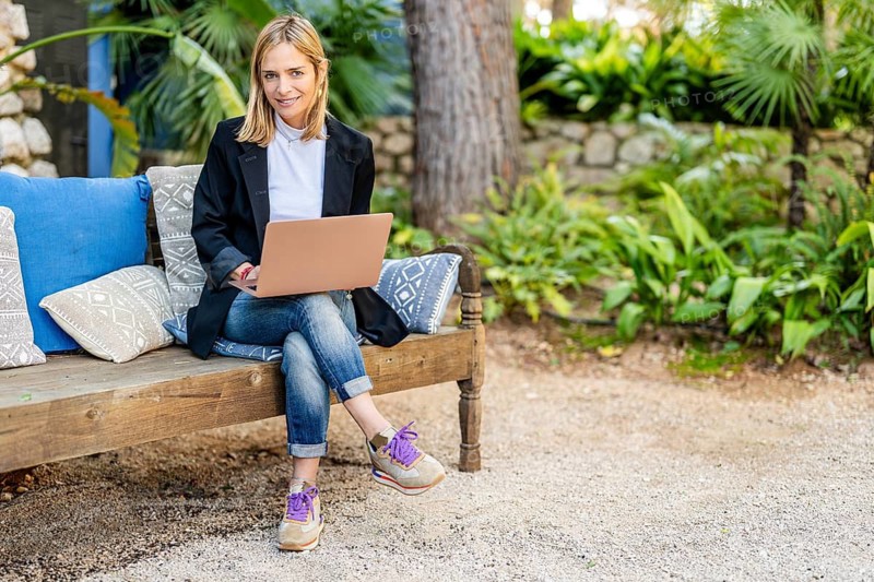 A woman sits on a wooden bench in a lush garden, working on her laptop Surrounded by greenery and soft cushions, she enjoys a tranquil outdoor work environment