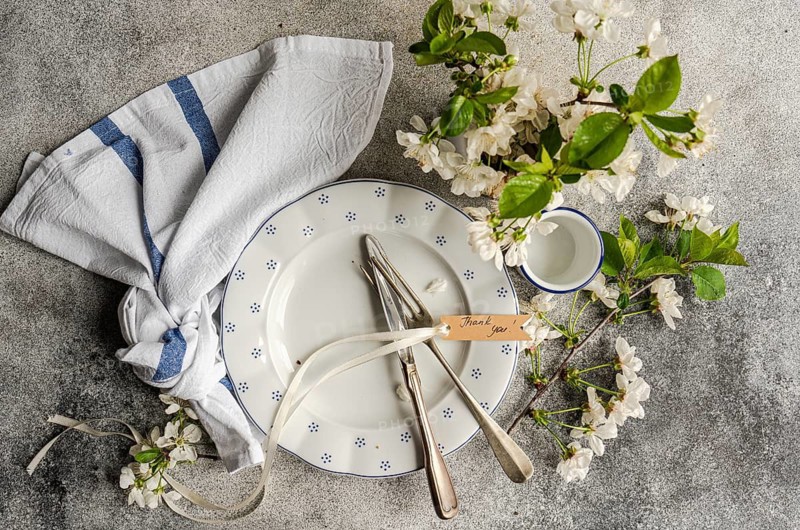A beautifully arranged table setting features a plate, cutlery, and a thank you note tied with ribbon. White blossoms and a folded napkin complete the elegant presentation