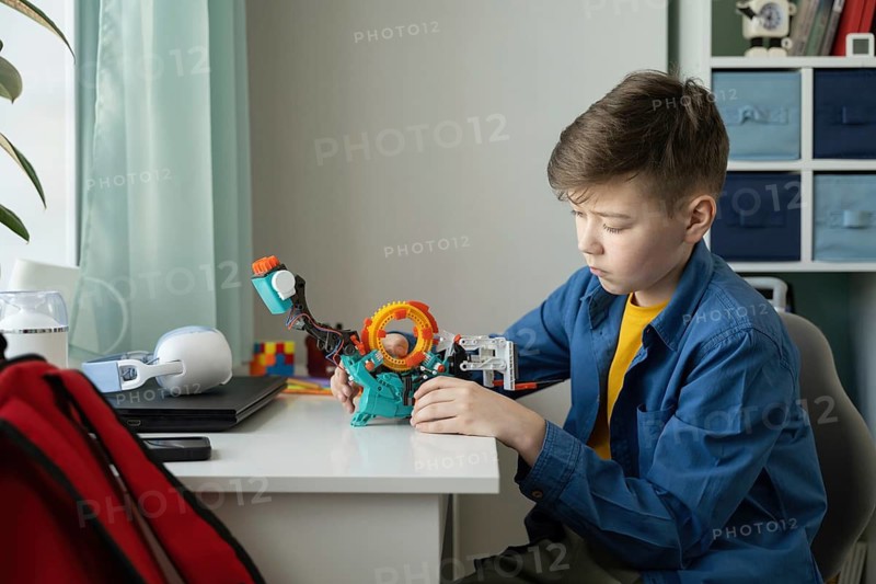 A young boy concentrates on building a complex toy structure with colorful blocks at a desk. The scene captures creativity, focus, and imaginative play in a cozy room setting