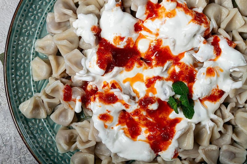 The dish features a generous serving of Turkish dumplings topped with velvety yogurt and a drizzle of spicy red sauce, displayed on a beautifully crafted plate with a green accent