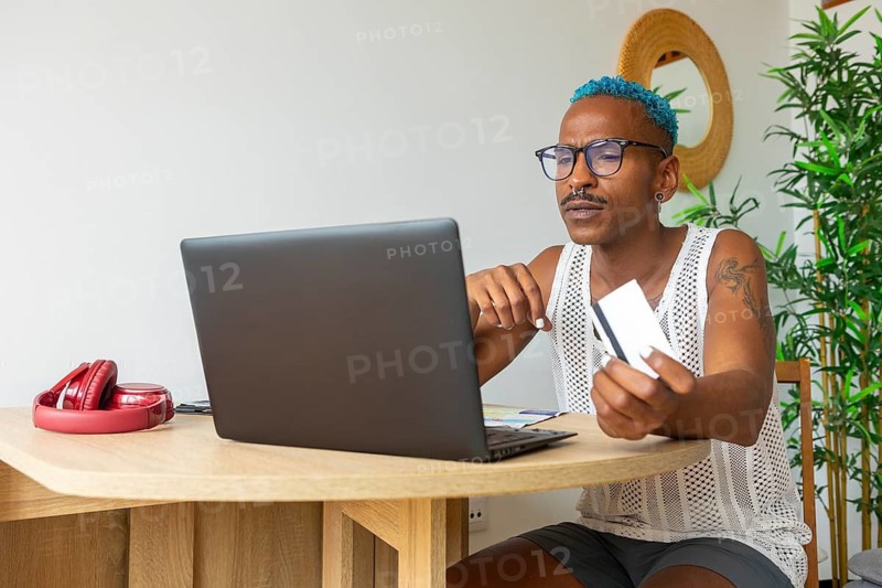 LGBTQIA+ man with blue hair using a laptop and credit card to prepare for holidays. A sense of pride and anticipation surrounds this festive moment