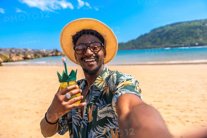 Young man soaking up the sun on a vibrant beach, sipping a refreshing cocktail and capturing joyful moments with a cheerful selfie