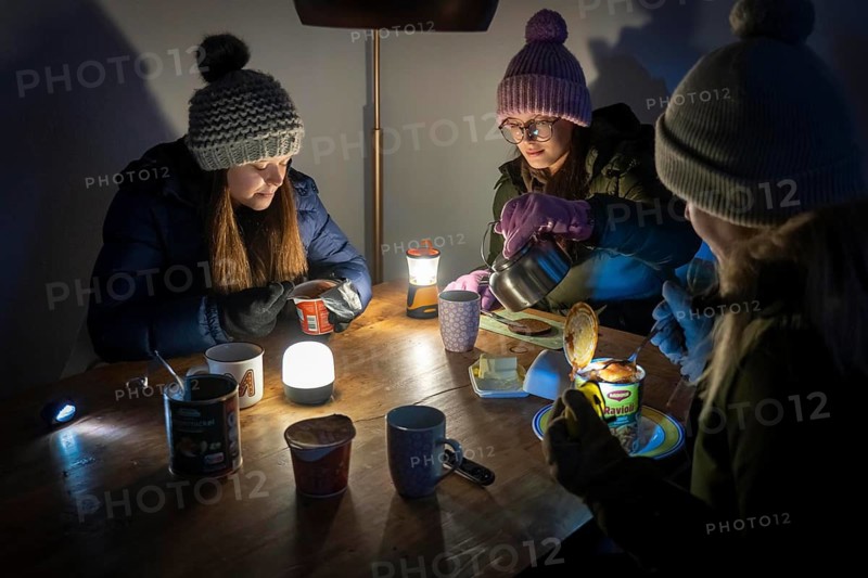 Symbolic blackout, extensive, prolonged power failure, in private surroundings, without electricity, heating, water, family at the dining table with warm winter clothes, sleeping bag, light from battery lights, flashlights, canned food, ready meals, heated on the camping stove, limited supply of food