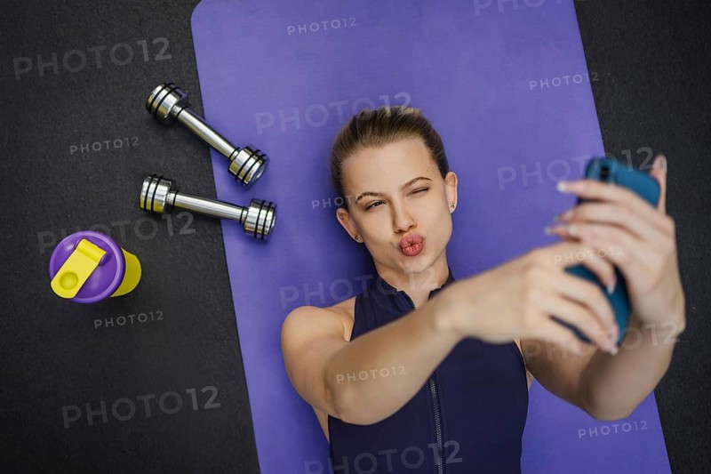 Blonde woman on purple yoga mat takes selfie after workout. Includes dumbbells and shaker on black surface. Showcasing fitness, fun, and self expression in a gym setting