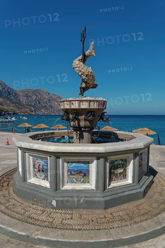 Decorative Neptune fountain OOlymbitissa with fish sculpture in front of the azure blue sea horizon of the coastal village of Diafani, Avlona, Diafani, Karpathos, Greece