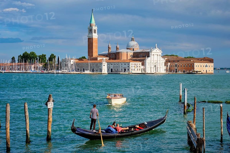 VENICE, ITALY, JUNE 27, 2018: Gondolier with tourists in gondola in lagoon of Venice by Saint Mark (San Marco) square with San Giorgio di Maggiore church in background in Venice, Italy