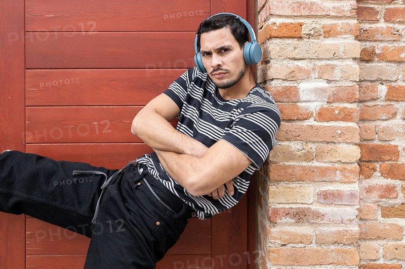 Young Cuban man in a striped shirt and headphones striking a dance pose against a brick wall. Captures a confident, casual vibe and showcases urban street style