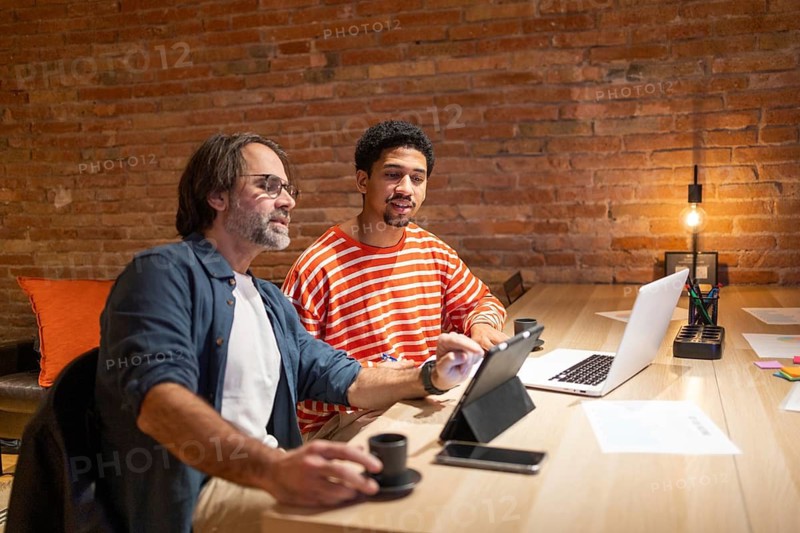 Two professionals in a coworking space engage in discussion, surrounded by laptops and office supplies, creating a productive environment with exposed brick walls