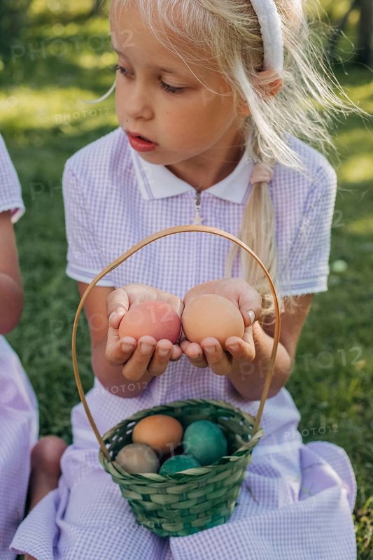 A young girl in a purple dress holds Easter eggs with a basket of more colorful eggs on her lap, sitting on grassy lawn. Sunlight enhances the playful scene