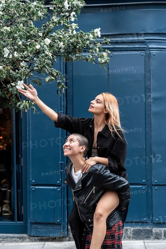 Joyful lesbian couple share a playful moment. One partner carries the other on her back to reach white blossoms against a blue wall, showcasing love and connection