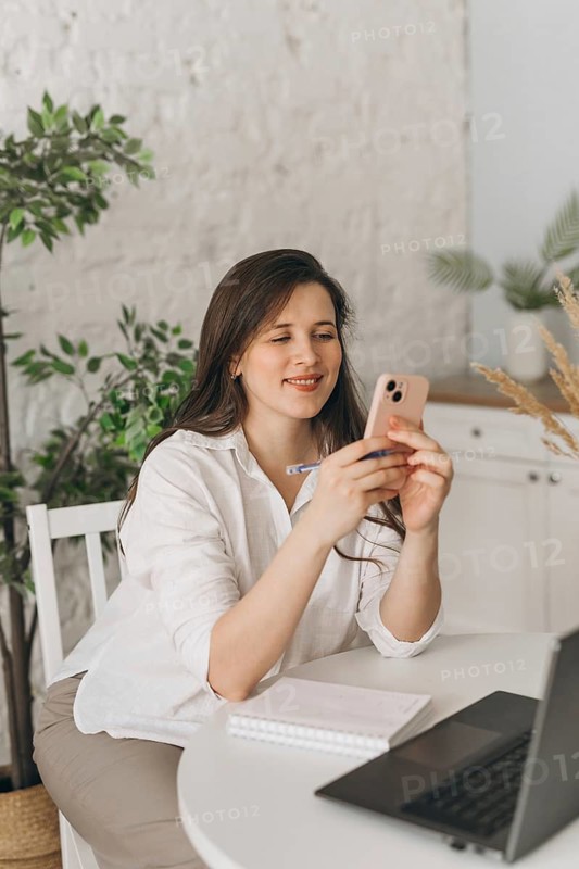 A nutritionist sits at a table with a laptop, phone, and notebook, engaging in a virtual consultation. Plants in the background create a calm and professional setting