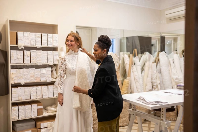 In a bridal atelier, a bride is assisted by a stylist as she selects her wedding gown. Fabrics and lace details surround them, creating a cozy, elegant atmosphere