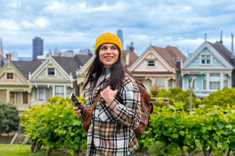 Woman traveler exploring alamo square while carrying a backpack and holding a smartphone, enjoying the urban cityscape and famous victorian architecture