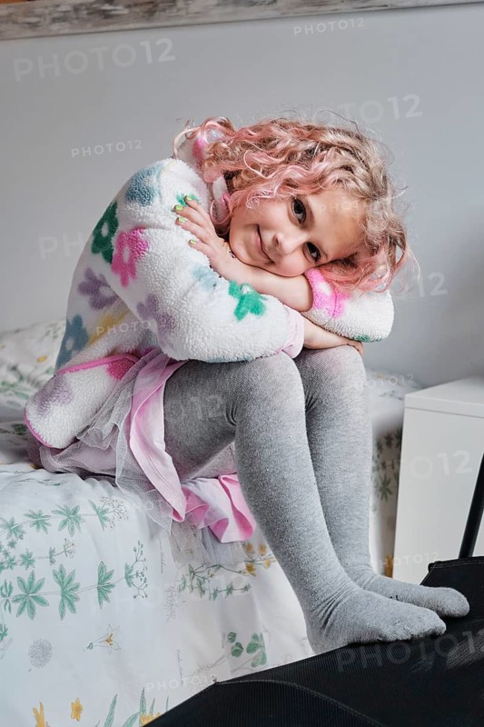 A young girl with curly hair sits on a bed, wearing a colorful sweater and gray leggings. She leans forward, resting her chin on her knees and smiling warmly