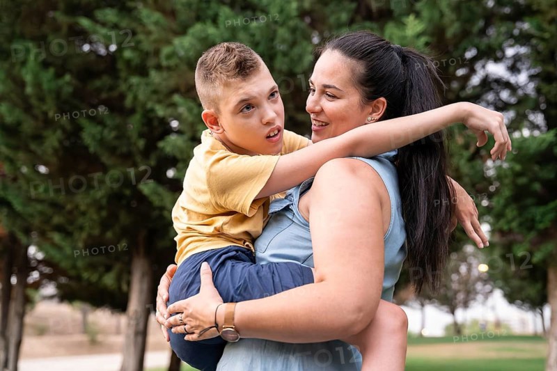 A loving mother holds her disabled son with cerebral palsy in a park, capturing a moment of joy and support amidst green trees, embodying familial bond and care