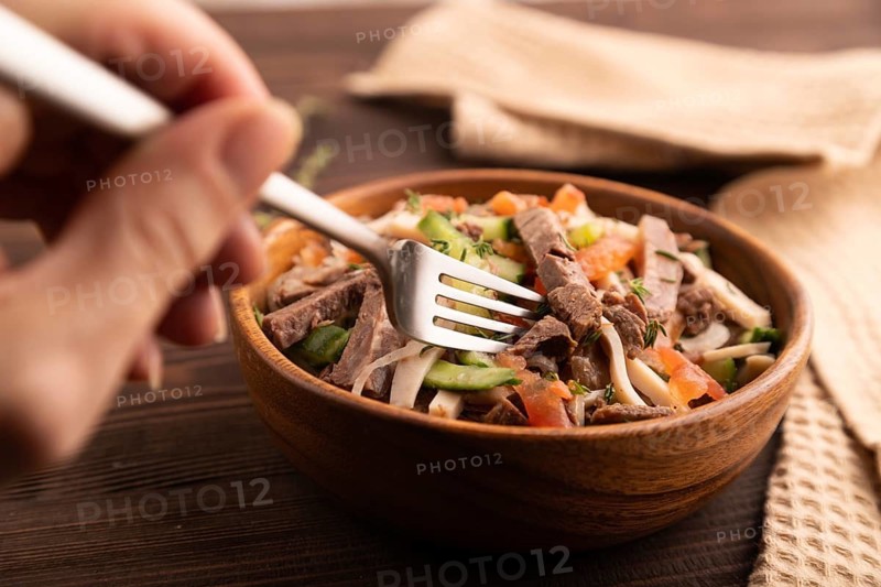Meat salad with beef and vegetables in wooden bowl with hand on brown wooden background and linen textile. side view, close up, selective focus