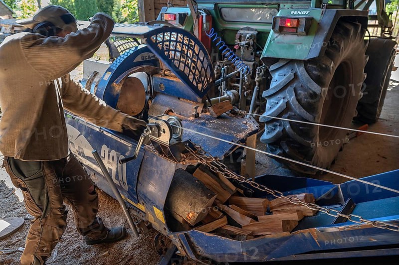Young man sawing and splitting beech logs with his saw cutting machine, powered by a tractor in a barn, Othenstorf, Meckleburg-Vorpommern, Germany