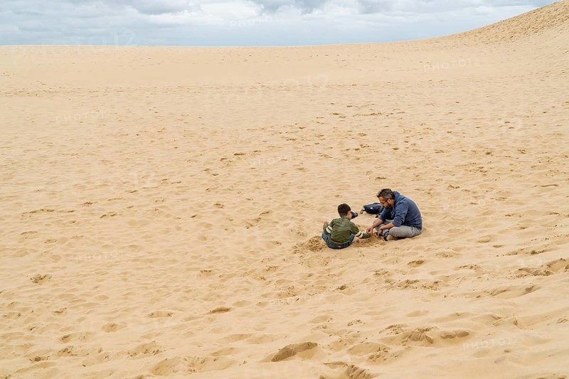 A father and son share a bonding moment, sitting on sand the dune of Pilat, under an overcast sky. The scene creates a sense of tranquility and connection