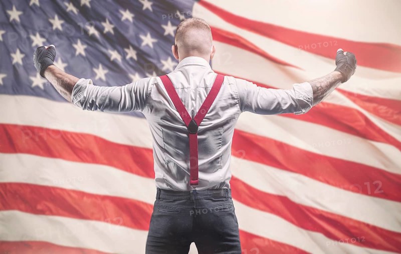 Image of a brutal man in a white shirt and red suspenders standing in front of the American flag. The concept of fist fights.
