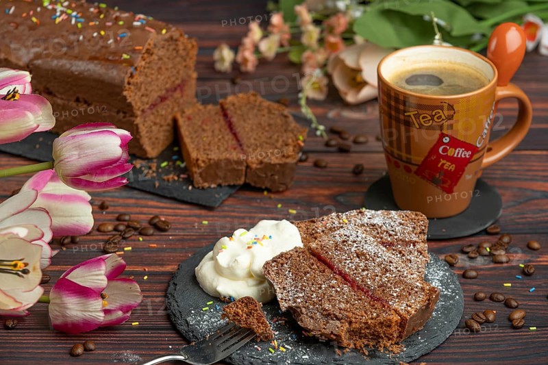 A slice of chocolate cake next to a cup of coffee decorated with tulips and whipped cream