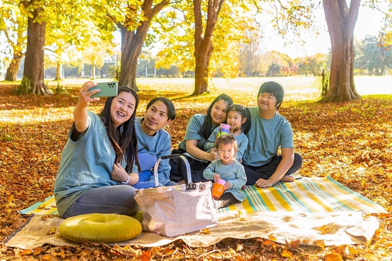 Happy Asian family having a joyful picnic outdoors, sitting on a blanket surrounded by autumn leaves. They're capturing the moment with a selfie, sharing smiles and laughter